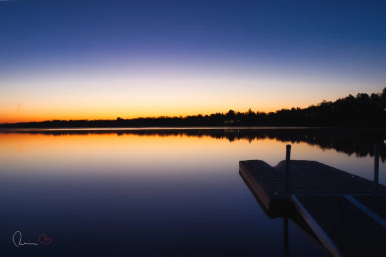 Early morning sunrise over calm water at Jacques-Cartier Park in Gatineau, Quebec, with a dock in the foreground and the tree line reflected on the lake.