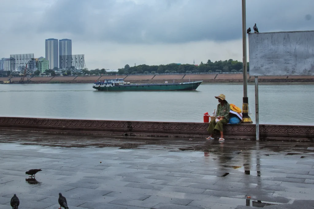 A rainy day along the riverside in Phnom Penh, Cambodia. A woman wearing a sun hat sits beside the promenade while a cargo boat passes on the water. Wet pavement reflects the cloudy sky, with pigeons in the foreground and city buildings across the river.