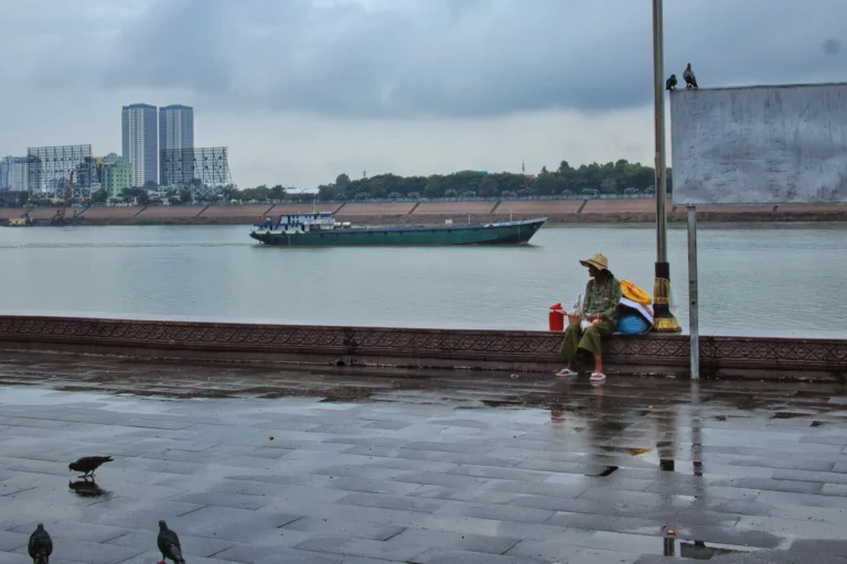 A rainy day along the riverside in Phnom Penh, Cambodia. A woman wearing a sun hat sits beside the promenade while a cargo boat passes on the water. Wet pavement reflects the cloudy sky, with pigeons in the foreground and city buildings across the river.