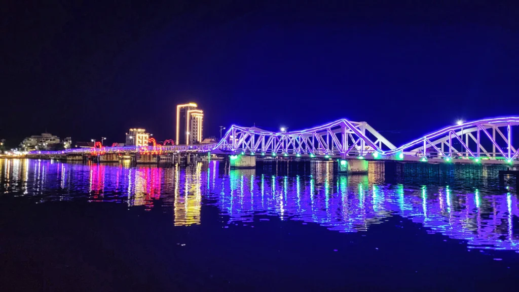 Illuminated bridge in Kampot, Cambodia, at night, with colorful lights reflecting across the river.